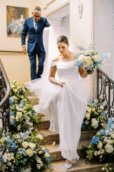 Bride descending a floral-lined staircase with her father, captured by a French wedding photographer.