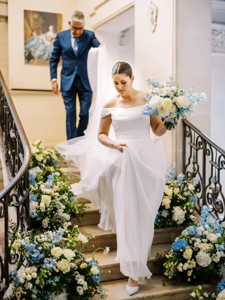 Bride descending a floral-lined staircase with her father, captured by a French wedding photographer.