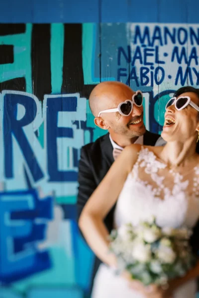 Laughing couple with heart sunglasses captured by a French wedding photographer against blue graffiti.