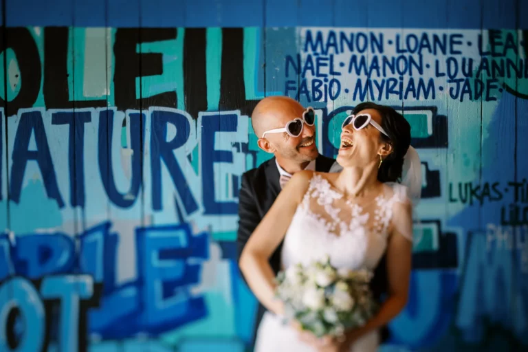 Laughing couple with heart sunglasses captured by a French wedding photographer against blue graffiti.