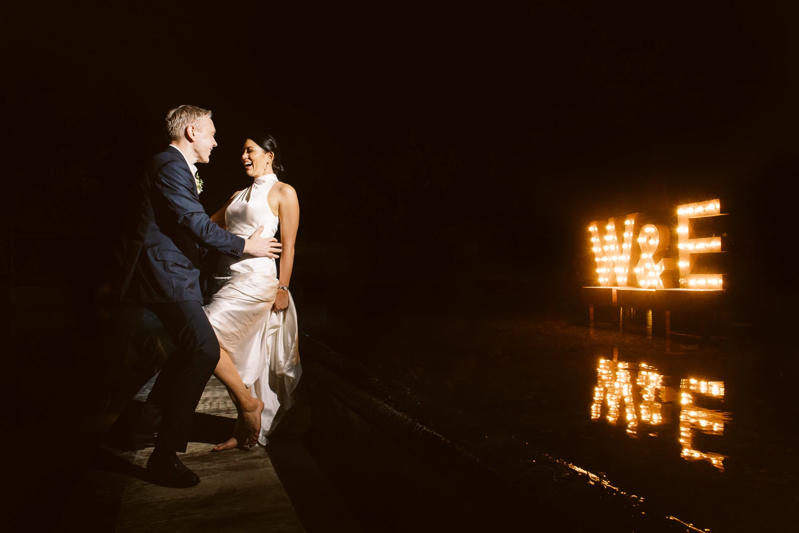 A Cannes wedding photographer captures a couple laughing by a luxury pool with glowing marquee letters at night.